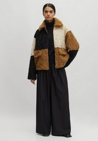 Texturierte, farbige Blockjacke in Schwarz, Creme und Braun, mit Kragen und frontalen Taschen, kombiniert mit weiten schwarzen Hosen.