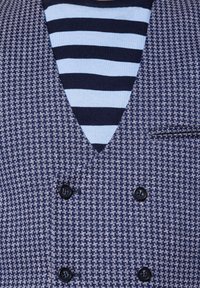 Granatowy kamizelka w wzór houndstooth z dwoma czarnymi guzikami, noszona na granatowej i jasnoniebieskiej koszuli w paski z dekoltem w serek.