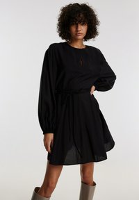 Schwarzes Kleid mit langen Ballonärmeln, Schlüssel-ausschnitt, tailliertem Bund, der mit einem Stoffgürtel gebunden ist, und einem fließenden Rock. Hergestellt aus leichtem Material.
