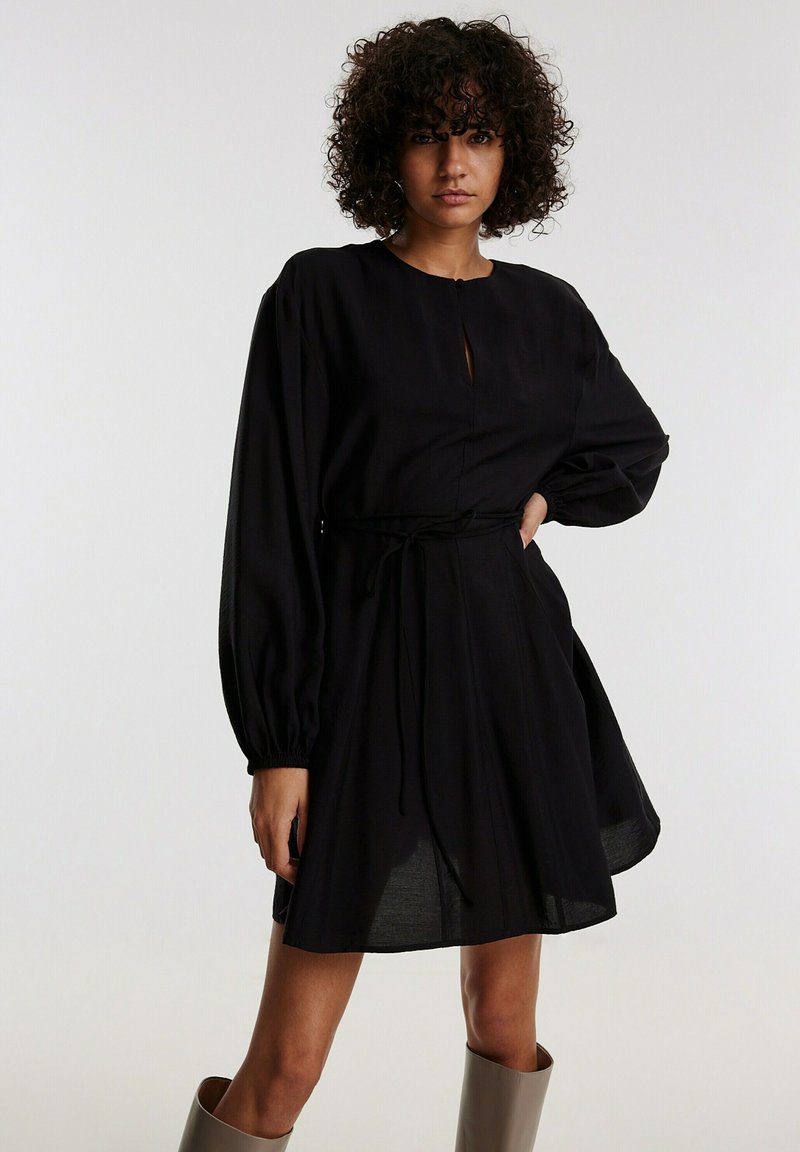 Schwarzes Kleid mit langen Ballonärmeln, Schlüssel-ausschnitt, tailliertem Bund, der mit einem Stoffgürtel gebunden ist, und einem fließenden Rock. Hergestellt aus leichtem Material.