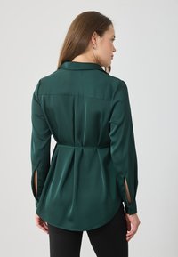 Camisa de cetim verde esmeralda com cintura ajustada, mangas longas com fendas laterais e bainha curva para uma silhueta descontraída.