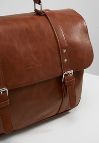 Sac cartable en cuir marron avec boucles argentées et logo "PIER.ONE" embossé sur le rabat avant sur fond uni.