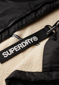 Capa exterior negra con cremallera sobre interior de forro polar crema. Presenta una prominente correa negra con el texto "SUPERDRY" y un clip de metal.