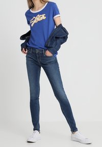 Camiseta azul con ribetes blancos y logo dorado de "Estrella", combinada con jeans ajustados oscuros y zapatillas blancas. Chaqueta de mezclilla sobre los hombros.