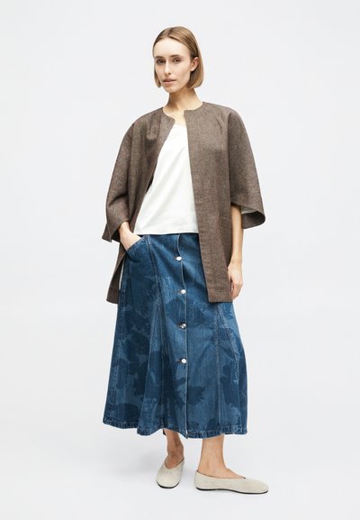 Γυναίκα που φορά καφέ κάπα με ανοιχτό μπροστινό μέρος, λευκή μπλούζα, μπλε τζιν φούστα με κουμπιά και μπεζ πλατφόρμες, στέκεται με το ένα χέρι στην τσέπη της φούστας.