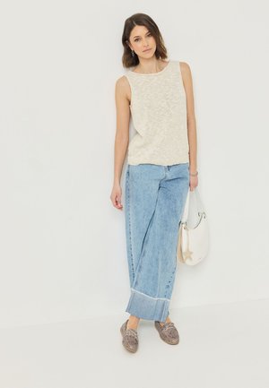 Kvinde lænende mod væggen iført ærmeløs beige top, blå jeans med vide ben og opsmøg, brune loafers, der holder hvid skuldertaske med stjernevedhæng.
