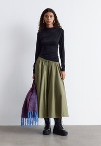 Schwarzes Langarmshirt mit geraffter Textur, olivgrüne weite Hose mit Kordelzug, schwarze Schnürstiefel und Tasche mit lila-blauen Akzenten.