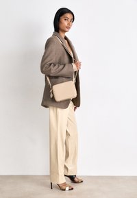 Borsa a tracolla beige con una texture liscia e chiusura con zip, indossata da una modella che indossa un cappotto marrone e pantaloni beige.