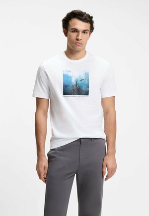 Junger Mann trägt ein weißes T-Shirt mit einem Grafikdruck von Haien unter Wasser und dunkelgraue Hose vor einfarbigem Hintergrund.