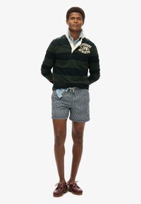 Camisa de manga larga a rayas negras y verdes, con jeans de mezclilla azul debajo, combinada con pantalones cortos a rayas y zapatos náuticos burdeos.