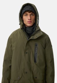 Olivgrüne wasserdichte Jacke mit Kapuze, Frontreißverschluss und Druckknöpfen. Verfügt über eine Brusttasche mit Reißverschluss und einen weichen, dunklen Strickschal.