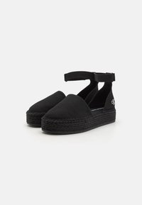 Chaussures espadrilles noires avec un matériau supérieur doux, une sangle à la cheville et une semelle en jute tissée. Présente un accent logo sur le côté.
