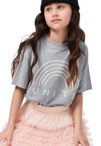 Djevojka s dugom tamnom kosom koja nosi crni beanie, sivi majicu s natpisom "UNITED" i duginim bojama, te ružičastu suknju s volanima.