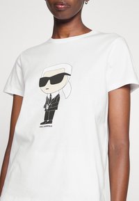 Balta īsām piedurknēm t-kreklu ar karikatūru attēlu, kurā redzams skaitlis melnā uzvalkā un saulesbrillēs, un zem tā ir teksts "KARL LAGERFELD".