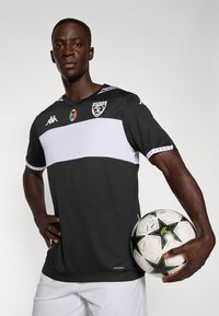 T-shirt de football noir et blanc à manches courtes avec logo, présentant une bande horizontale blanche sur la poitrine. Le joueur tient un ballon à motifs.