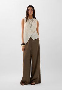 Leichtes, cremfarbenes, ärmelloses Oberteil mit strukturiertem, gekreuztem Design, kombiniert mit lockeren, braun gemusterten Hosen und flachen Sandalen.