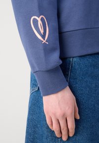 Sweatshirt bleu avec un poignet côtelé, présentant un design de cœur brodé rose sur la manche gauche. Porté sur un tissu en denim.