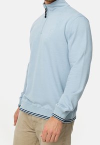 Sweat à capuche bleu clair avec une encolure zippée au quart, des poignets et un ourlet côtelés avec des accents rayés foncés. Texture douce, branding minimal.