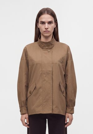 Femme aux cheveux bruns et lisses porte une veste ample beige avec un col montant et des poches boutonnées, debout devant un fond clair uni.