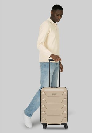 Man in beige shirt met lange mouwen en spijkerbroek loopt terwijl hij een beige harde koffer met vier wielen en een telescopisch handvat trekt.