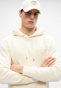 Krem sweatshirt s prednjim džepom, kapicom koja se podešava i GANT logotipom. Uparen s odgovarajućom kapom s izvezenim tekstom. Glatka tekstura.