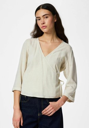 Femme aux longs cheveux foncés portant un chemisier beige à manches trois-quarts et un jean bleu foncé, debout devant un fond blanc.