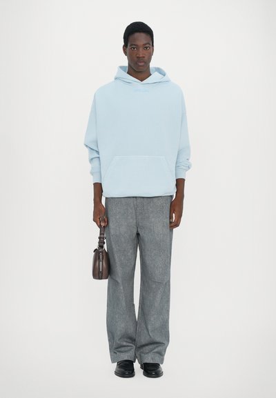Felpa oversized blu chiaro con tasca frontale, abbinata a pantaloni grigi a gamba larga. Il modello tiene una piccola borsa a tracolla marrone scuro. Scarpe nere.