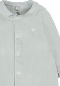 Maglietta per bambino di colore verde chiaro con righe, colletto rotondo, chiusura a bottoni e un delicato motivo a cuore sul petto. Tessuto morbido e testurizzato.