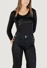 Maglietta nera a maniche lunghe con logo "Guess" in strass, abbinata a jeans scuri a vita alta con dettagli in bottone argentati.