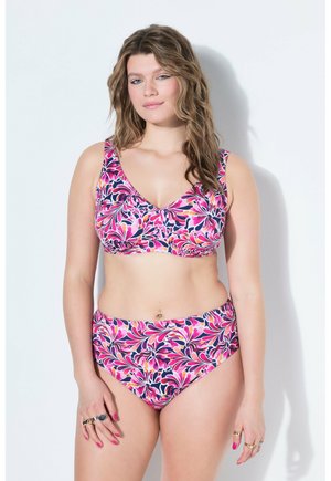 Frau trägt einen bunten pinken, violetten und orangen Blumenbikini mit Ringen an den Fingern, steht vor einem schlichten weißen Hintergrund.