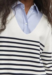 Femme portant un pull à col en V blanc avec des rayures horizontales noires sur une chemise bleu clair boutonnée, montrant le haut du torse et le cou.