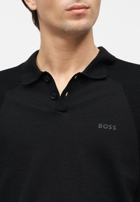 Νεαρό άτομο που φορά μαύρο πουκάμισο με γιακά, δύο κουμπιά και μικρό λογότυπο BOSS στο αριστερό μέρος του στήθους.