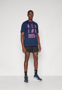 Mørkeblå bomulds T-shirt med pink grafisk tekst "BERLIN 2025," parret med sorte shorts og mørkeblå sneakers med hvide striber.