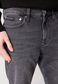 Sive traperice s prilagođenim dizajnom, s Calvin Klein logotipom na džepu, suptilnim šavovima i metalnim detaljima.