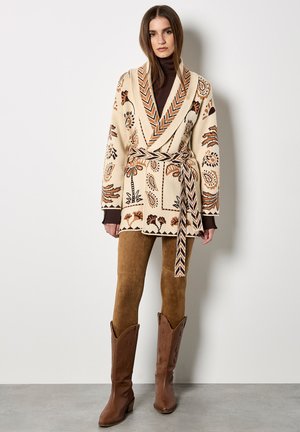 Cárdigan beige con patrón, cuello de chal y cinturón para atar, que presenta diseños florales en tonos marrón, naranja y negro. Combinado con pantalones de ante marrones y botas de cowboys.