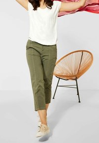 Olivgröna cropped byxor med sidofickor och sömdetaljer, kombinerat med en vit t-shirt med kort ärm och ljusa sneakers. Håret utsläppt.