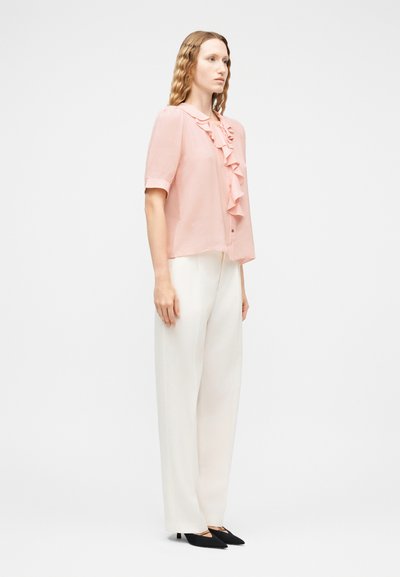 Femme debout portant un chemisier à manches courtes rose clair avec des volants, un pantalon blanc à jambes larges et des talons pointus noirs contre un fond uni.