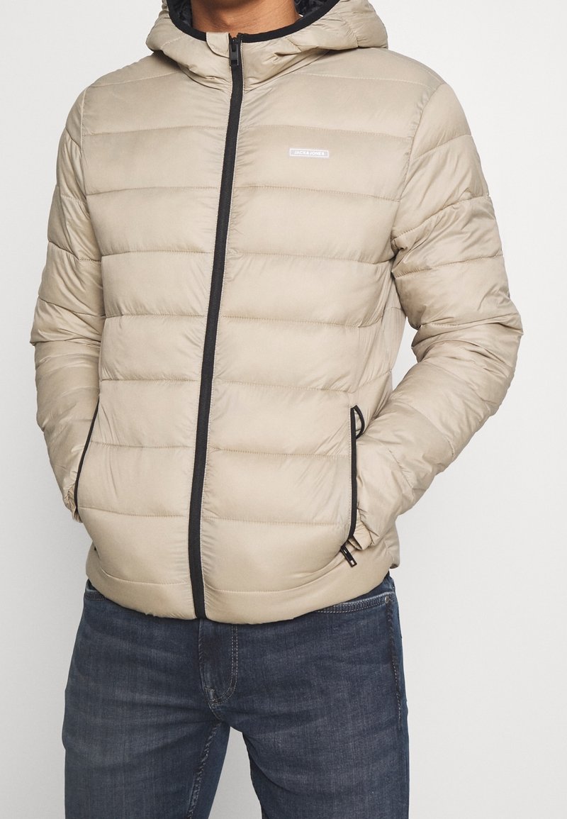Beżowa kurtka puchowa z kapturem, z pikowanym wzorem, czarnym zamkiem błyskawicznym i bocznymi kieszeniami. Lekka tkanina dodaje teksturę i ciepło.