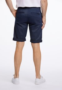 Pantaloni corti blu navy con orlo arrotolato e una texture in cotone liscio, dotati di due tasche posteriori e indossati con sneakers bianche.