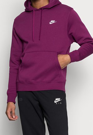 Osoba ubrana w fioletową bluzę Nike z kapturem i kieszenią z przodu oraz czarne spodnie Nike Air, z jedną ręką w kieszeni, na jednolitym tle.