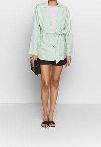 Veste blazer en lin vert menthe légère avec une ceinture à la taille, manches longues et une poche avant unique, portée avec un short noir et des sandales.