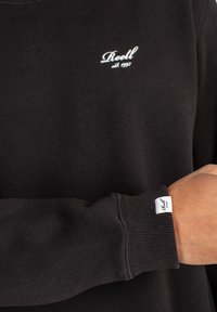 Sudadera negra de algodón, con un logo blanco "Reell" en el pecho y una etiqueta de marca en el puño con textura acanalada en las mangas.