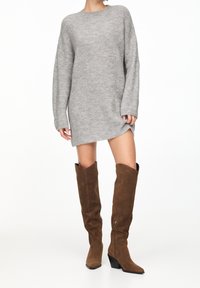 Robe-pull grise en tricot avec manches longues, tissu texturé et fente latérale. Bottes hautes jusqu'aux genoux en daim marron avec talons inclinés et fermetures éclair.