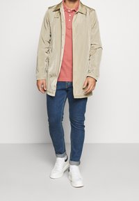 Ljusbeige vattentät kappa med krage, knappstängning och sidofickor, bärs över en rosa pikétröja och blå åtsittande jeans med vita sneakers.