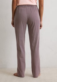 Vrouw die blootsvoets staat, gekleed in roze geometrisch patroon met elastische taillebroek en een lichtroze top, gezien vanuit de rug.