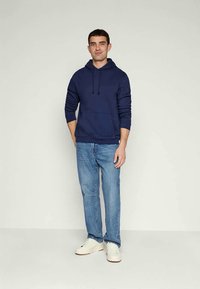 Young man standing, wearing a navy hoodie, blue jeans, and white sneakers against a plain light background.