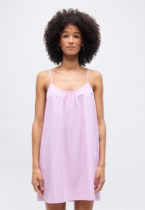 Kobieta o kręconych włosach, ubrana w różowo-białą sukienkę w paski bez rękawów z cienkimi ramiączkami, stojąca na tle jasnego, jednolitego tła.