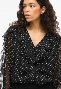 Blusa nera a maniche lunghe con pois bianchi, collo arricciato, maniche traslucide e chiusura con bottoni. Tessuto morbido.