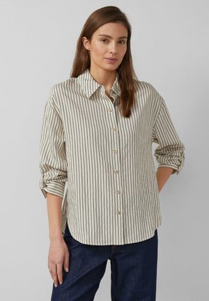 Femme portant une chemise beige à boutons avec des rayures noires verticales et des manches retroussées, associée à un pantalon bleu foncé, debout devant un fond gris.
