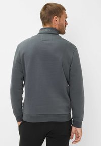 Felpa grigia con colletto alto e polsini a costine. Presenta un branding sottile sul colletto e una texture liscia. Decorata con tasche laterali.